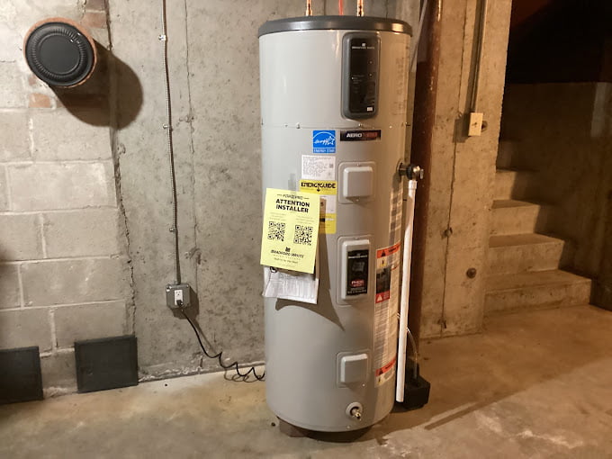 In the basement, a tall white water heater stands near the concrete wall and stairs, its yellow instruction tag catching the light. For expert assistance with such systems, consider reaching out to Eric Sabo Plumbing Heating & Cooling for all your plumbing needs.