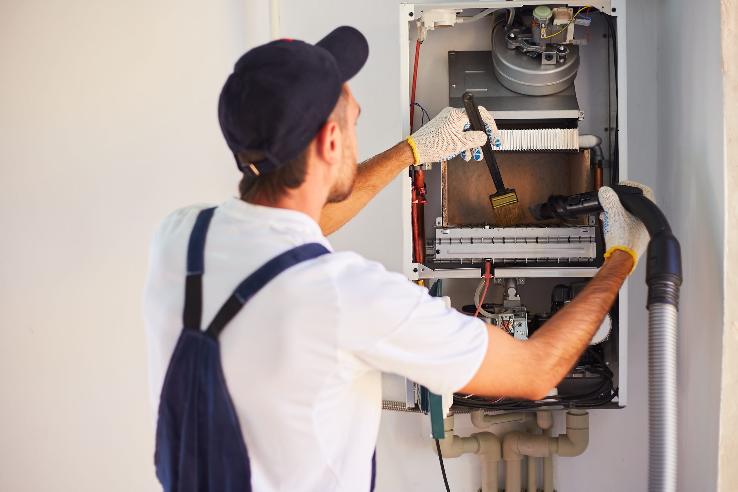 A technician in overalls and a cap is skillfully repairing a wall-mounted heating appliance with precision tools and protective gloves.