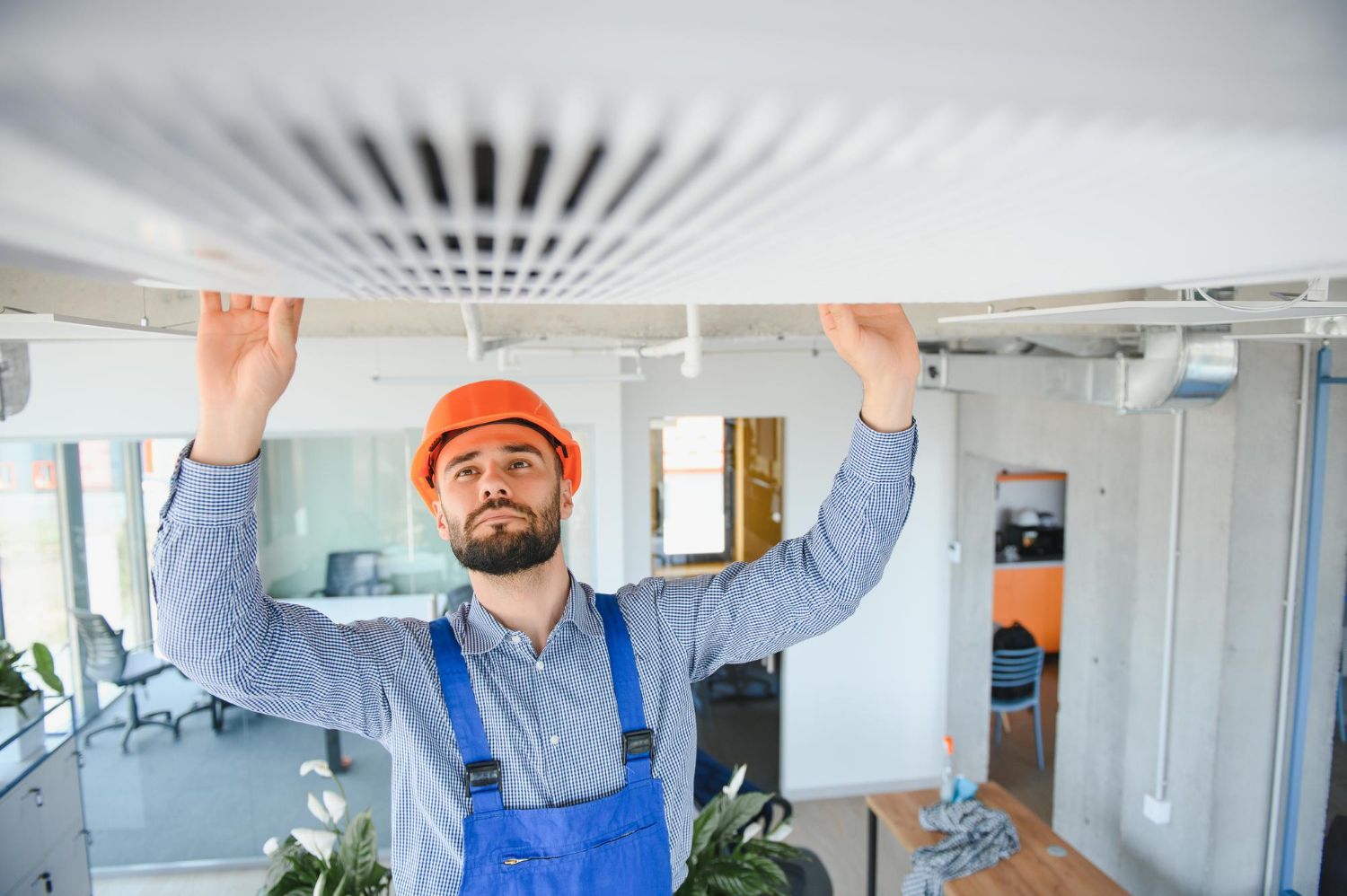 Man in hard hat and overalls inspecting or fixing an air conditioning unit on a ceiling in an office.