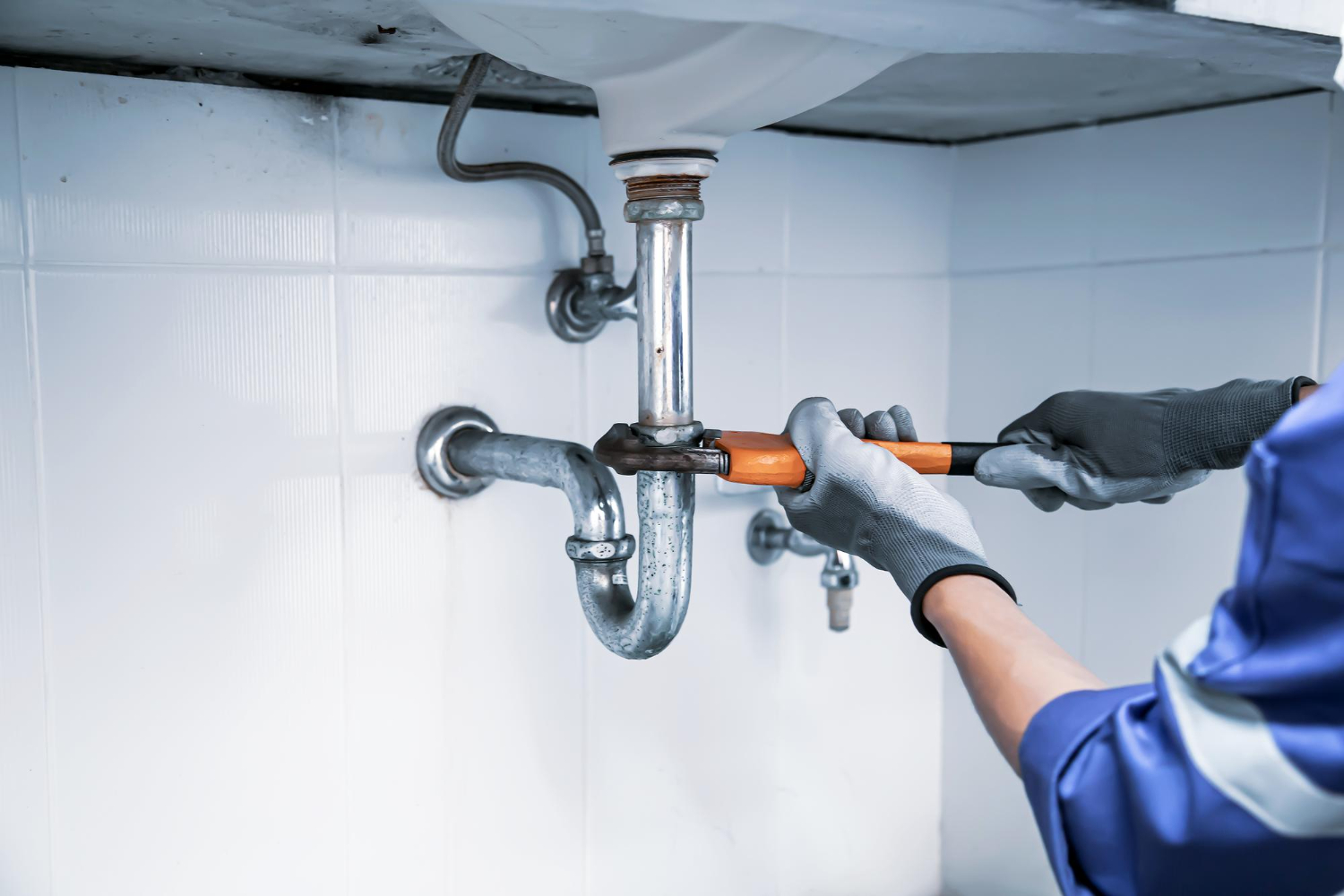 A plumber wearing gloves fixes pipes under a sink with a wrench.