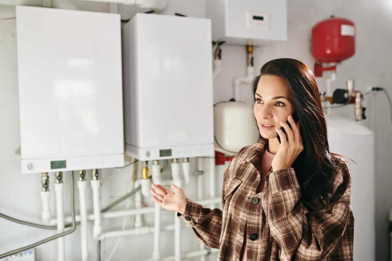 Woman talking on phone, standing in a boiler room with heating equipment and pipes in the background.